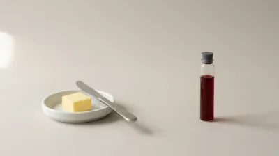 A single pat of butter on a white dish beside a small glass vial of blood-red liquid on a pale grey surface in soft directional daylight
